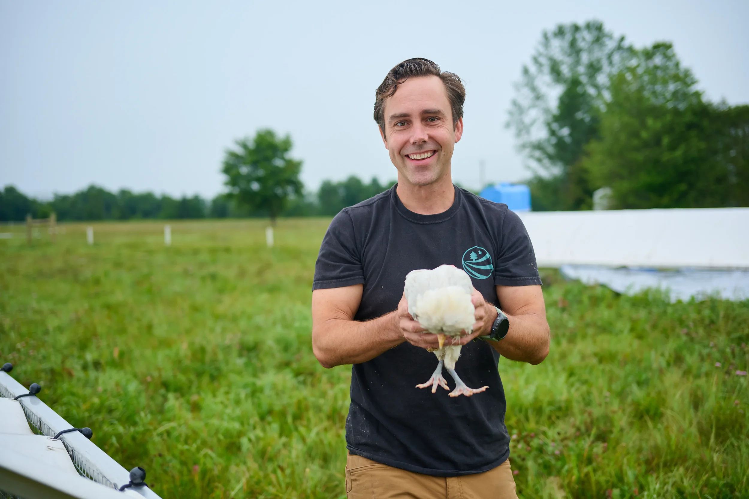 A smiling man in a black T-shirt holding a white chicken outdoors in a grassy field with trees in the background.