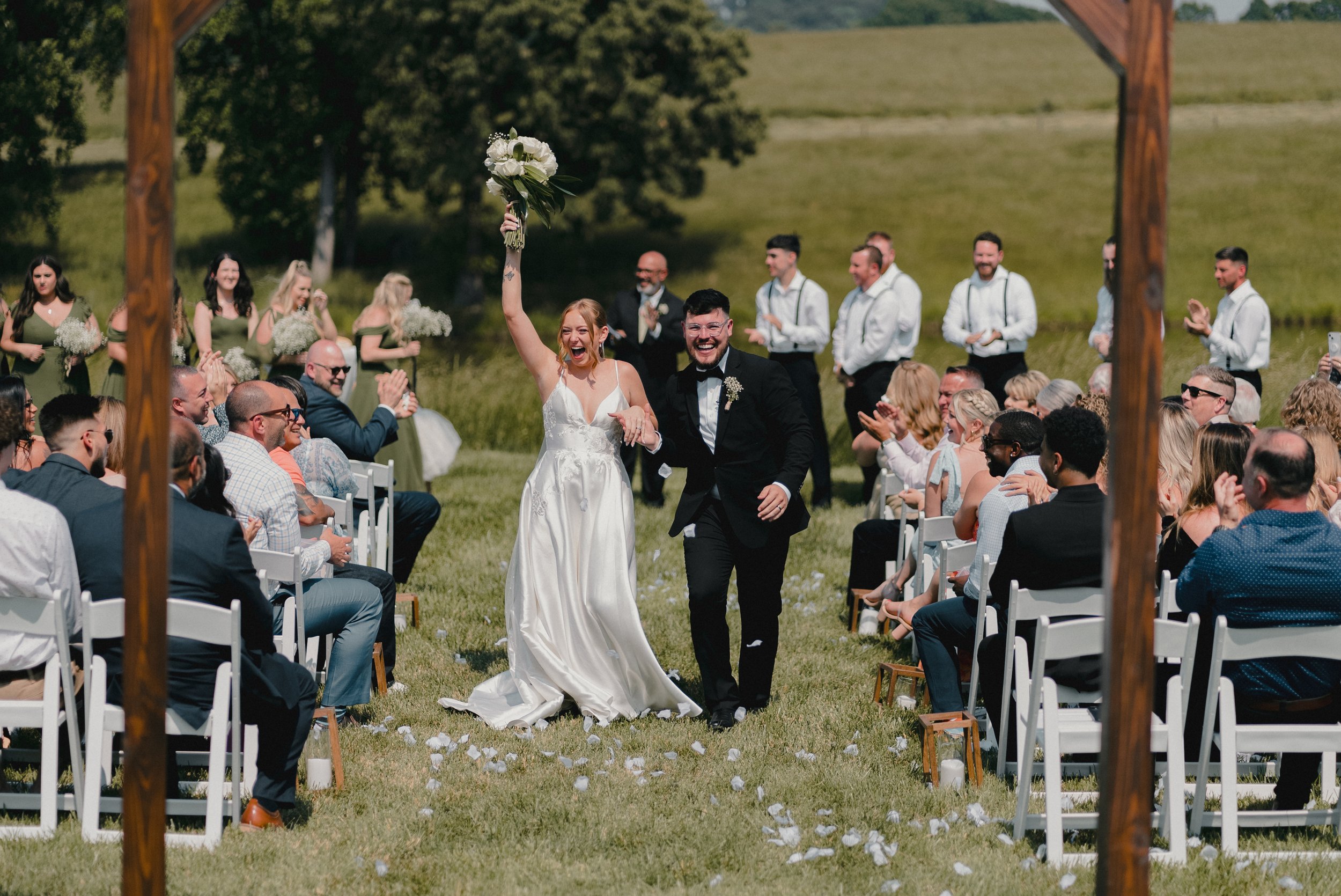 A newlywed couple walking down the aisle celebrating after their wedding ceremony outdoors, with the bride holding a bouquet in the air and everyone clapping and smiling around them.