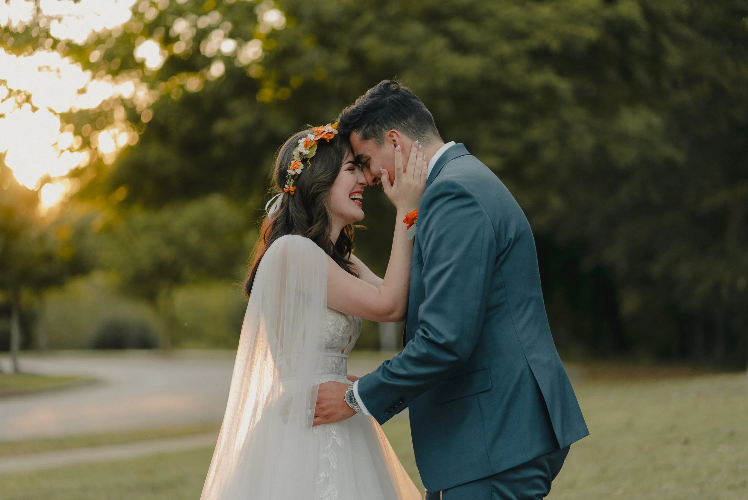 A bride and groom embracing outdoors at sunset, touching foreheads with smiles, the bride wearing a white wedding dress and floral headband, the groom in a teal suit.
