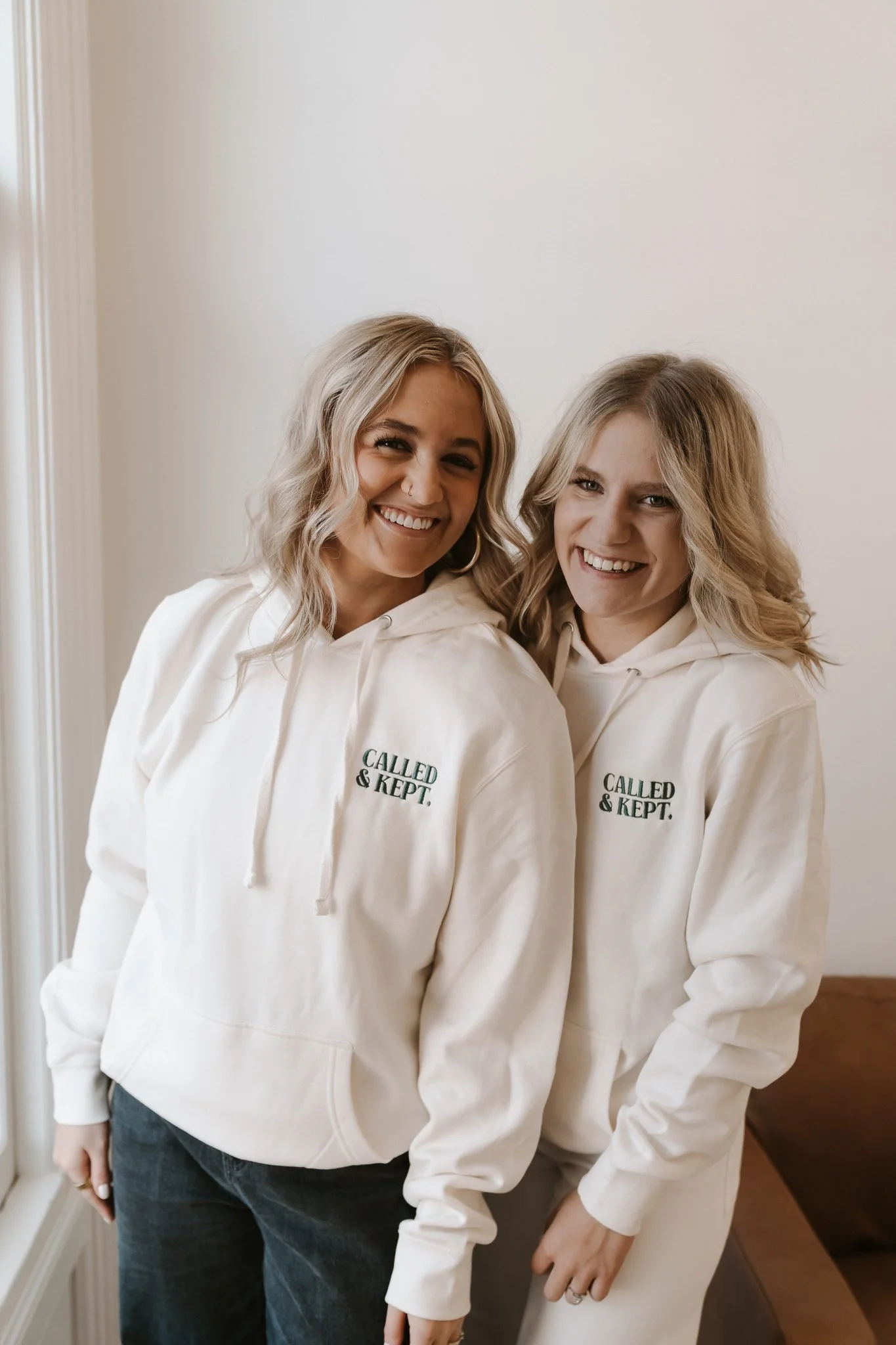 Two women smiling and wearing matching white hoodies that say "CALLED & KEPT." standing indoors near a window and a brown couch.