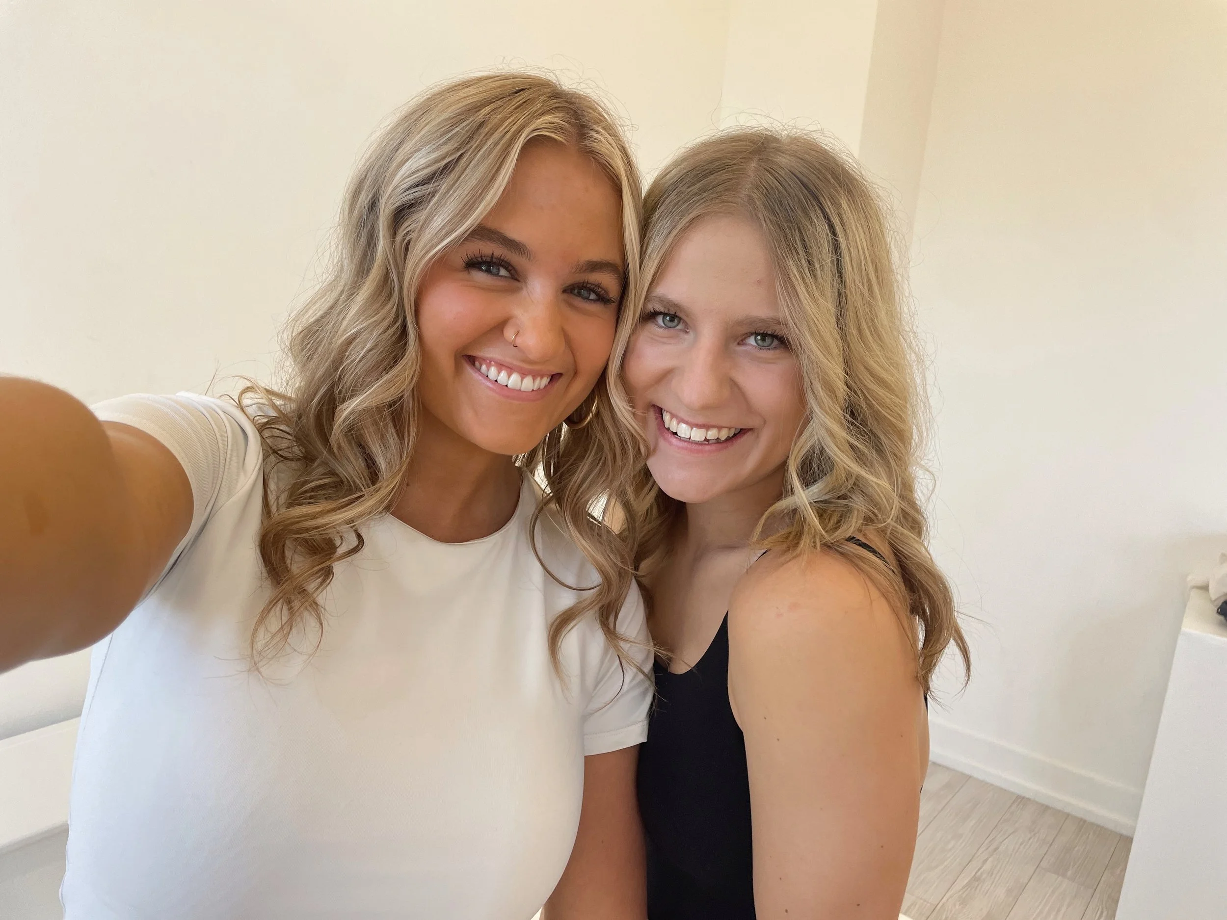 Two women smiling and taking a selfie in a well-lit room, one wearing a white shirt and the other in a black top, with neutral-colored walls in the background.