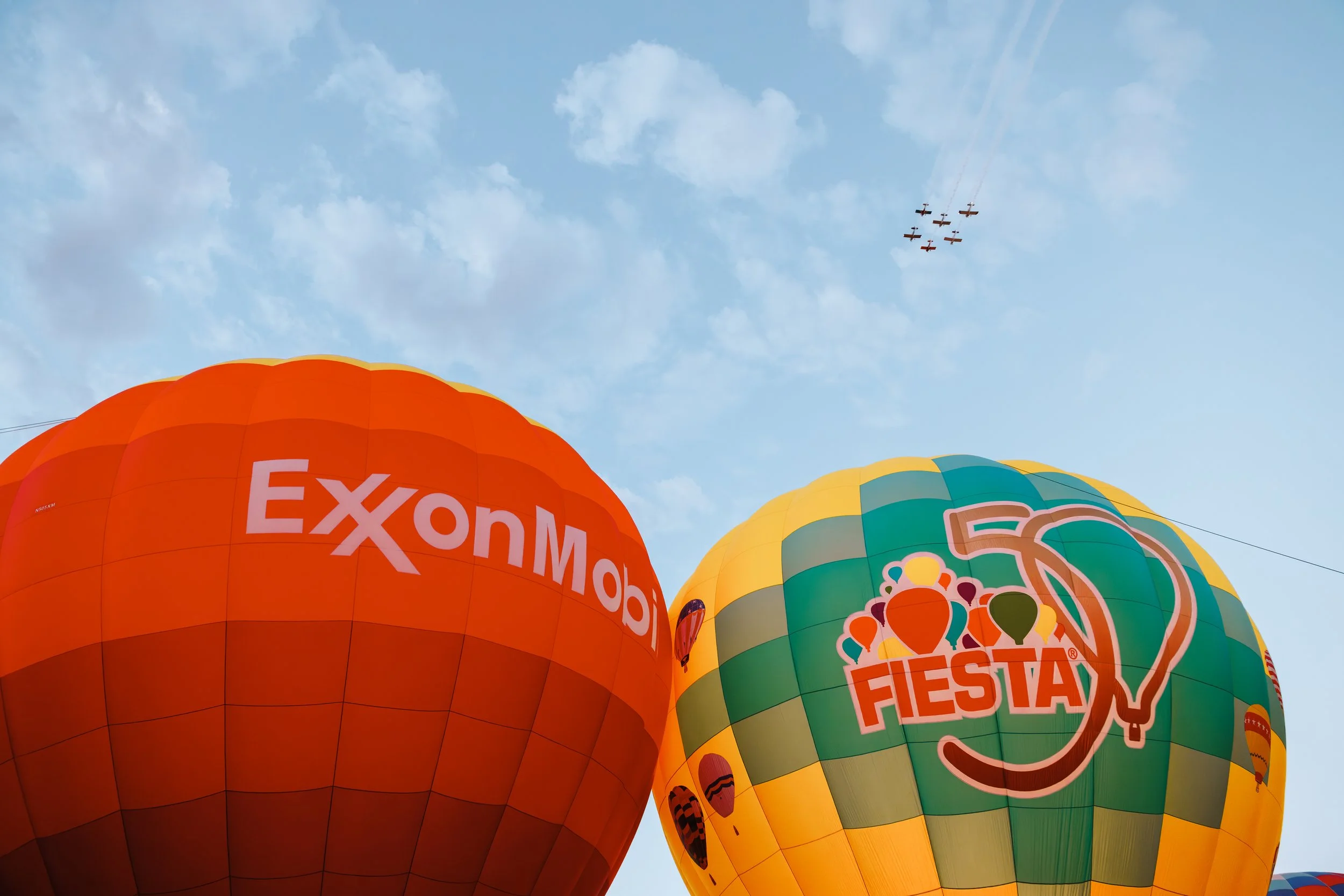 Two large hot air balloons against a blue sky, airplans in the distance,one orange with ExxonMobil branding and one yellow and green with a 50th Fiesta anniversary logo.