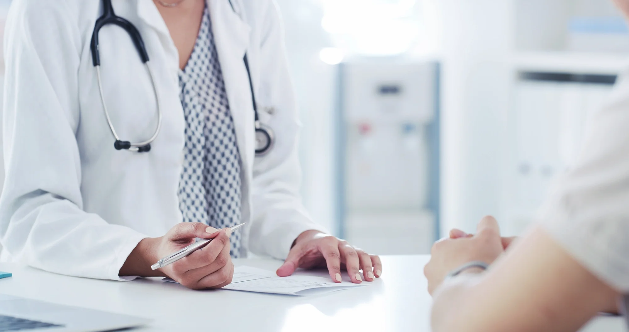 A healthcare professional, possibly a doctor, holds a pen and appears to be explaining or discussing something with a patient in a medical setting with a stethoscope around her neck.