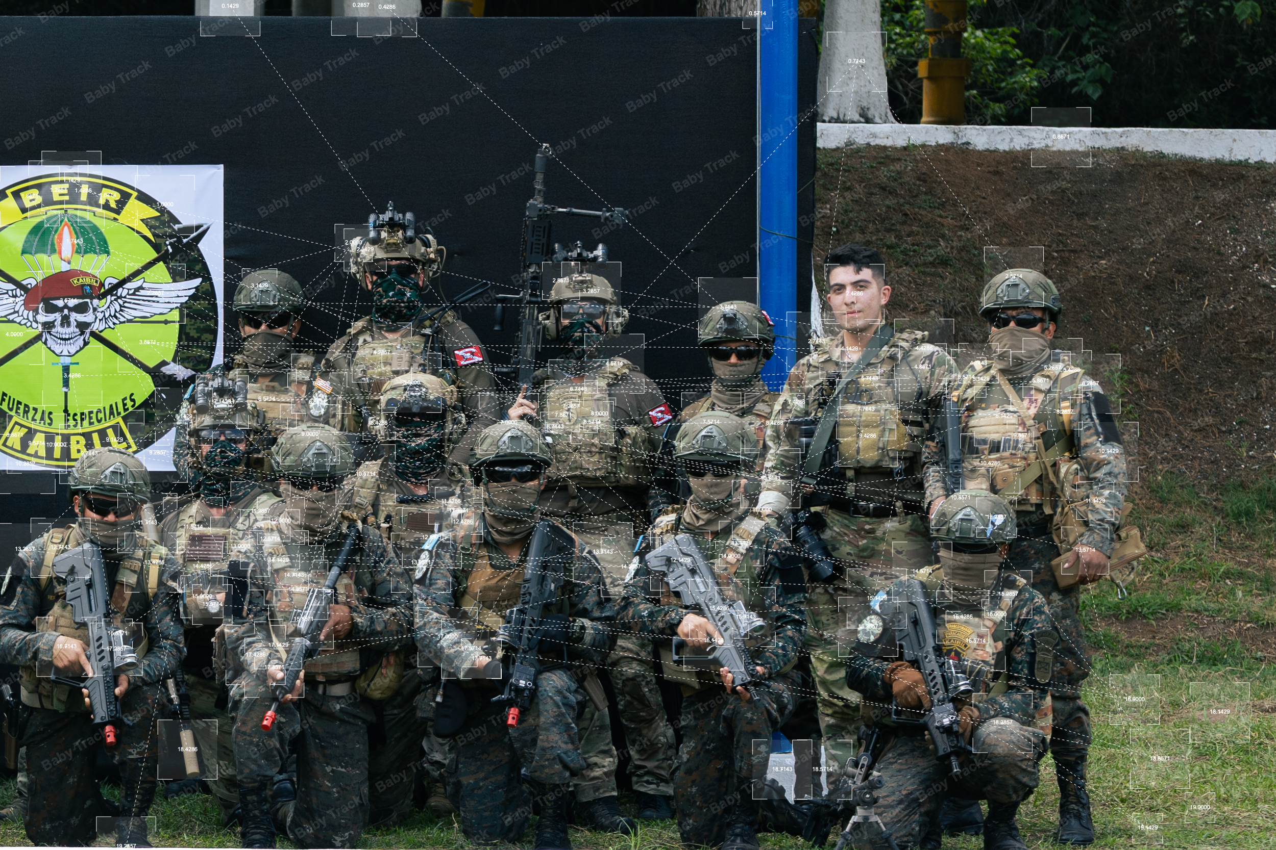 Group of military personnel in camouflage uniforms and tactical gear, holding firearms, standing outdoors on grass. Some are wearing helmets and sunglasses. Behind them is a black banner with a skull and wings logo, and a sign with the words "Baby Track" and a skull emblem. The setting appears to be a training or demonstration area.