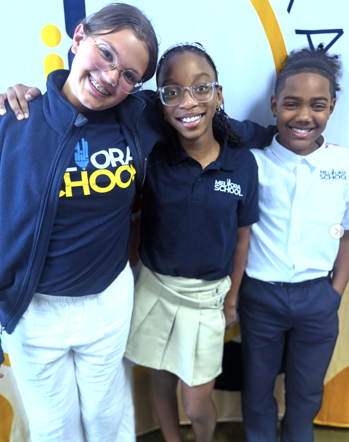 Three students standing together, smiling, with their arms around each other, in front of a colorful background at Meliora School.