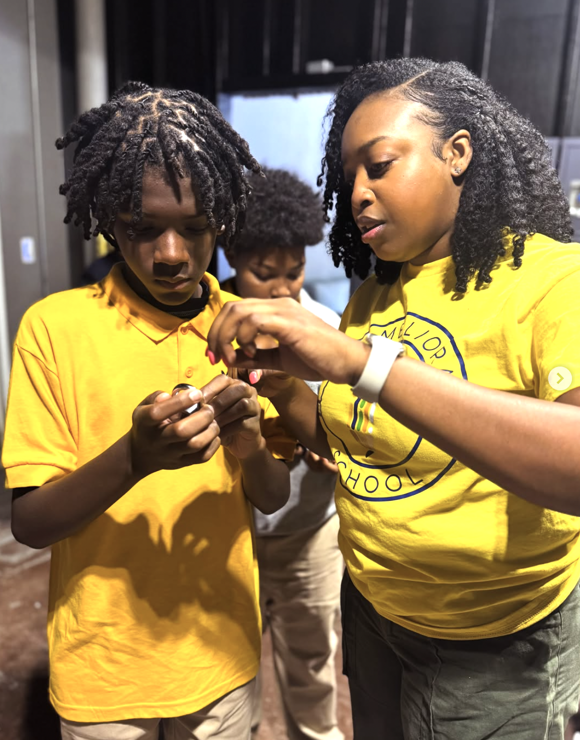 A woman and a young man, both wearing yellow t-shirts, are looking at a small object in their hands together. Another person in the background, also wearing a yellow shirt, is visible.