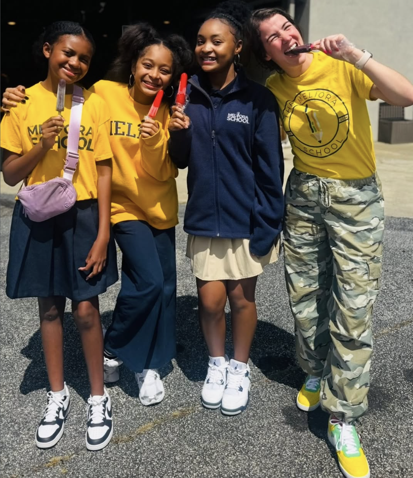 Four young girls standing outdoors, smiling, and holding ice pops. They are dressed casually, two in yellow Meliora School t-shirts, one in a navy jacket, and one in camouflage pants and colorful sneakers.