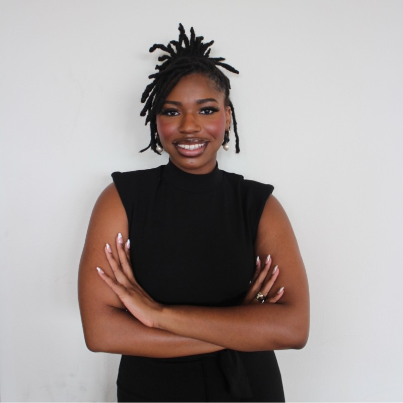 A woman with short dreadlocks wearing a sleeveless black top, smiling with her arms crossed against a plain white wall.