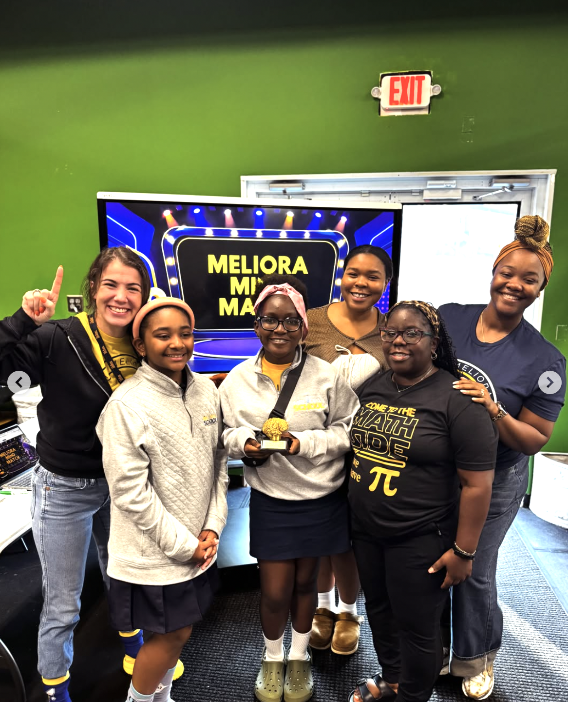 Group of six women and girls smiling in a room with green wall, standing in front of a TV displaying a game show titled 'Meliora Match'. One girl in the center holds a trophy. All are dressed casually, some wearing glasses, and appear happy.