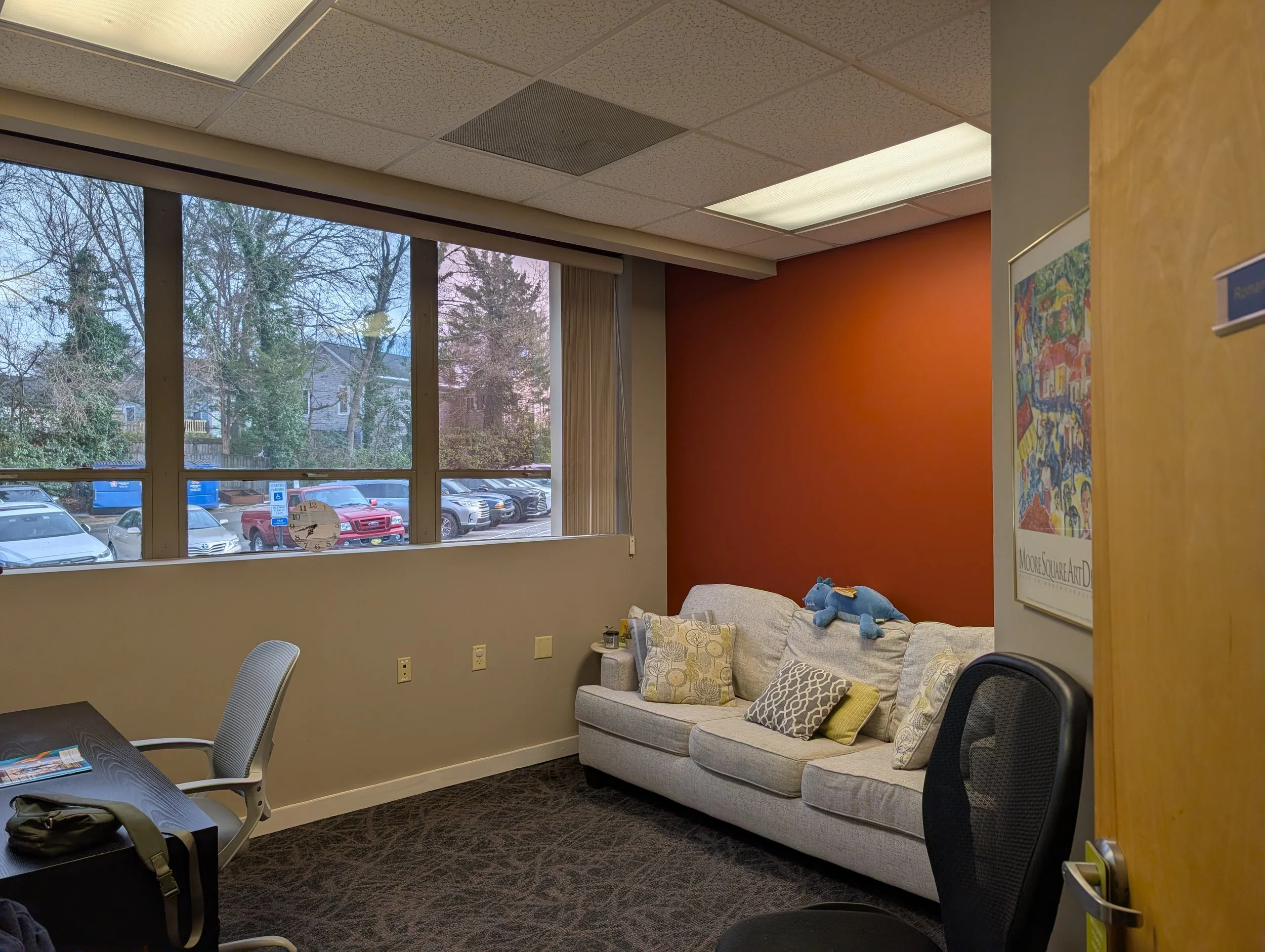 Downtown Raleigh Office interior with a window showing parked cars and trees outside, a beige sofa with pillows and a blue stuffed animal, a small table, chairs, and wall art.