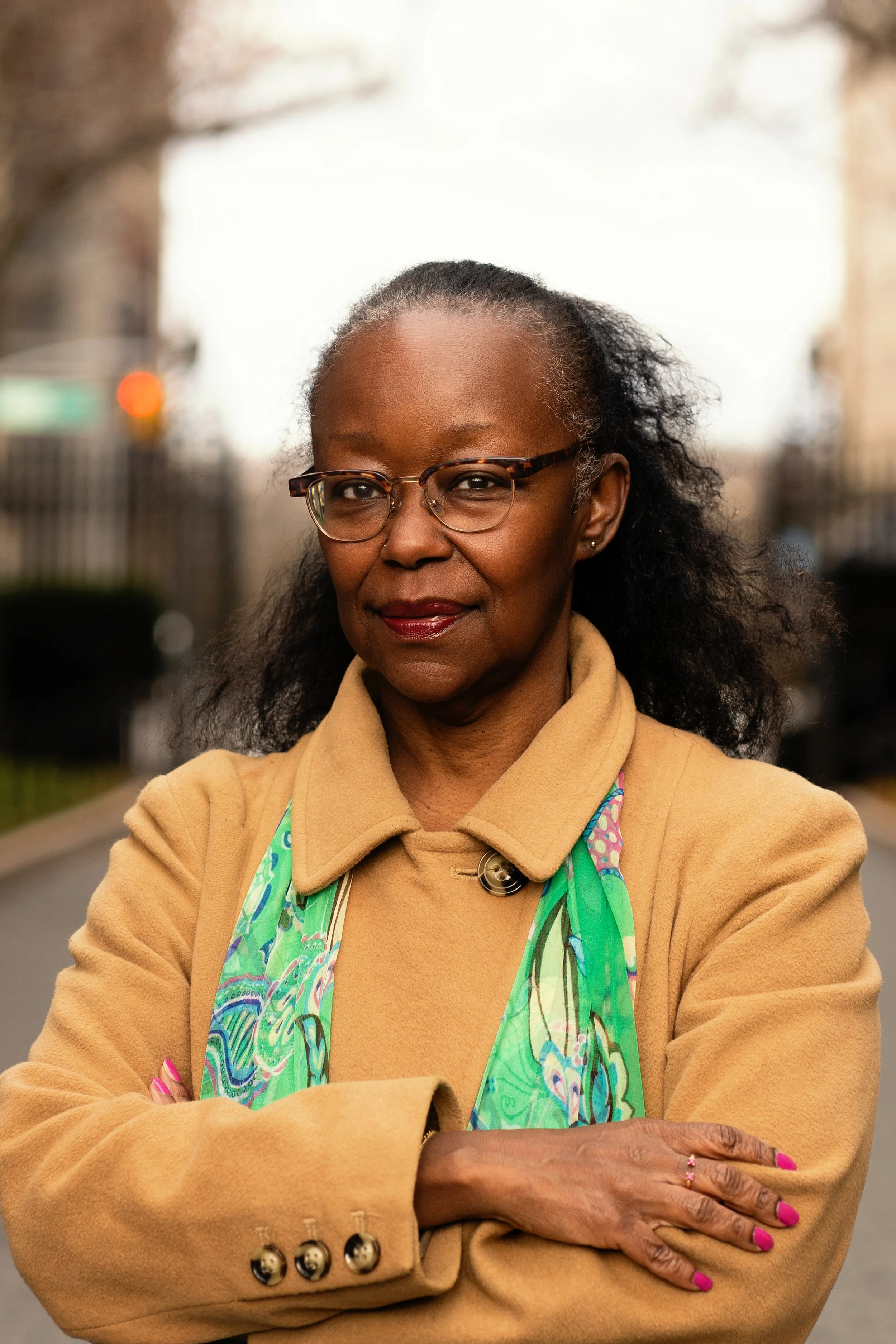 A confident middle-aged woman with glasses, wearing a tan coat, colorful scarf, and pink nail polish, crossing her arms outdoors.