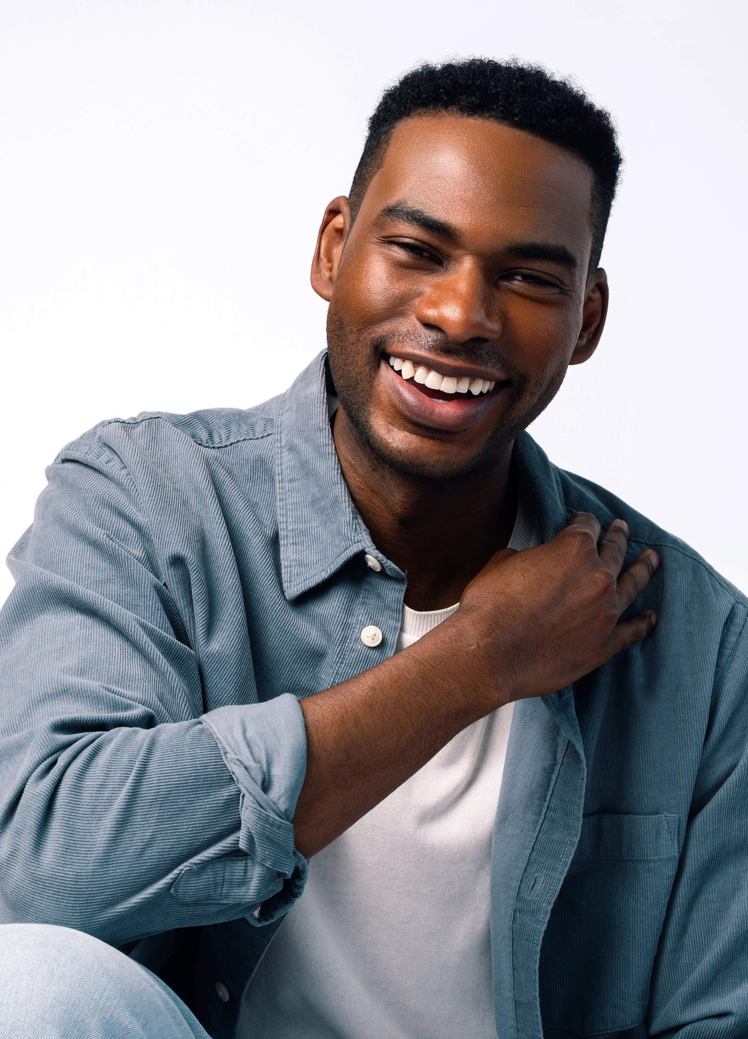 A headshot of a  young man with dark skin, short curly black hair, and a wide smile, wearing a light blue button-up shirt over a white t-shirt, touching his shoulder with his right hand and looking at the camera against a plain white background.