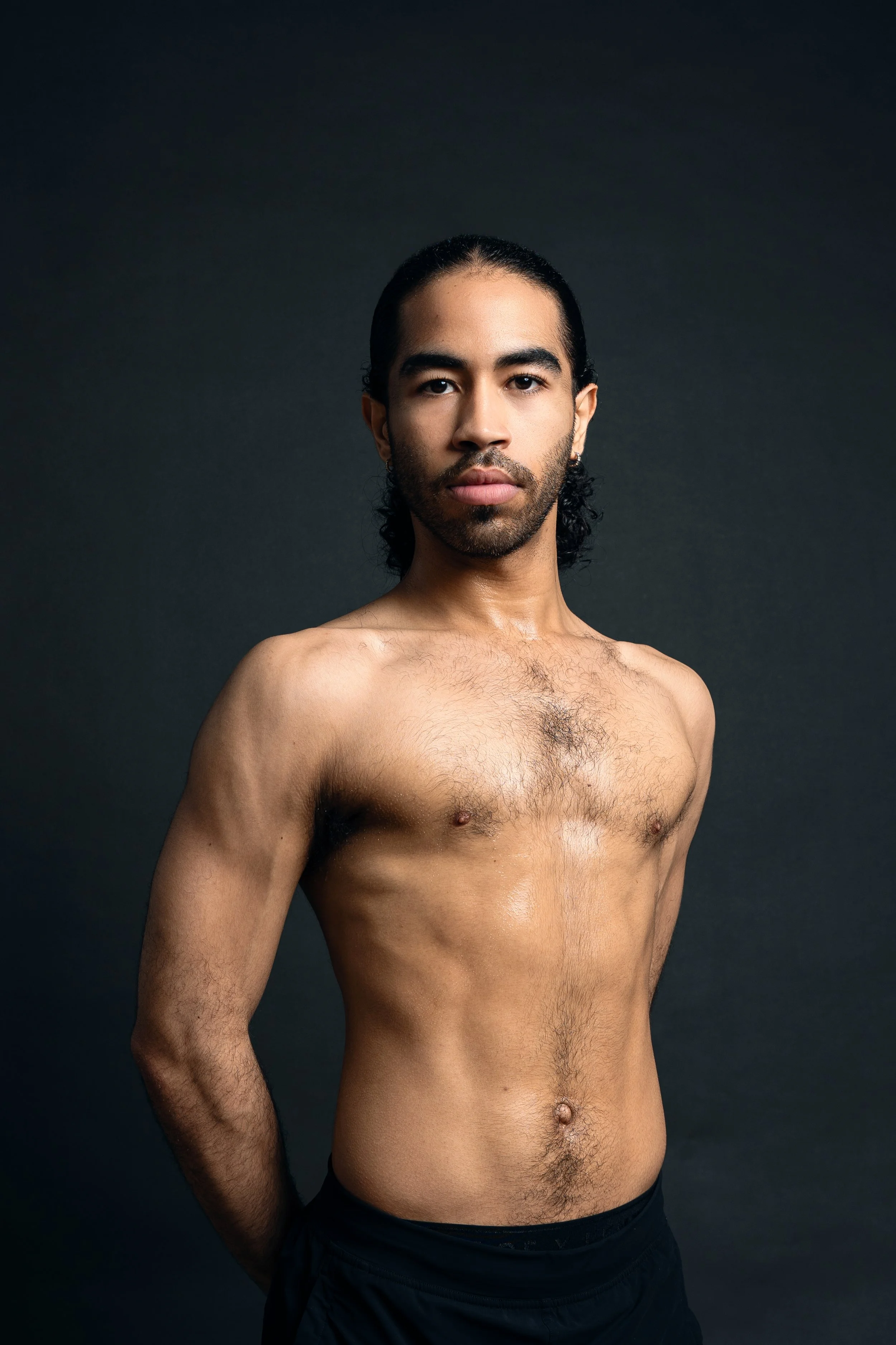 Portrait of a shirtless man with long dark hair, standing against a dark background.