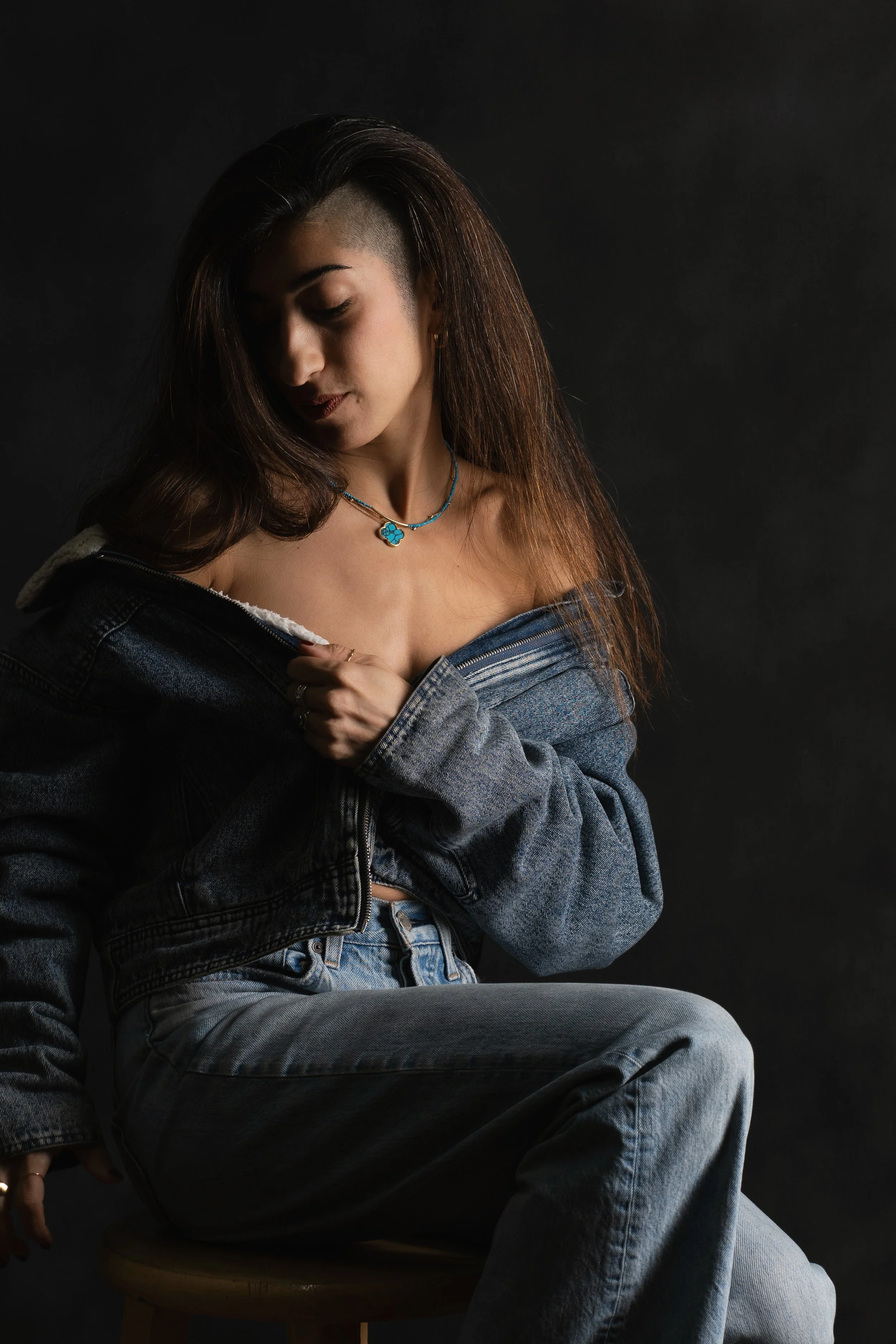 A portrait of a woman with long dark hair and a partial undercut, wearing a denim jacket off her shoulders and jeans, sitting on a stool against a dark background, looking down.