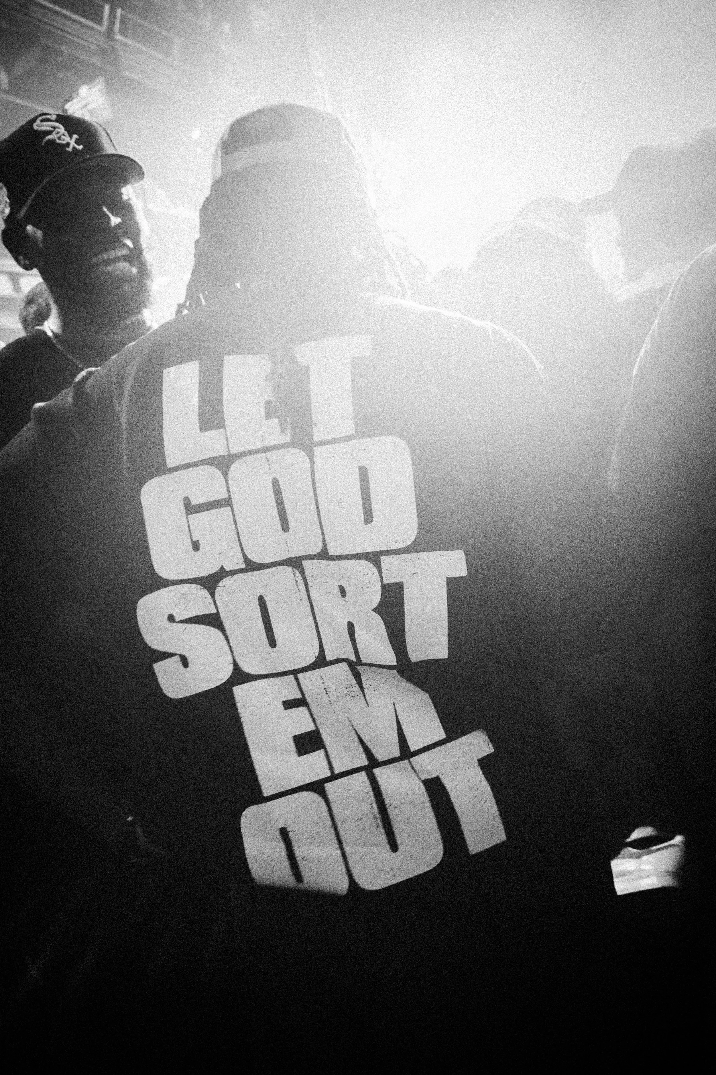 Back of a person wearing a sweatshirt that says "Let God Sort Em Out" while in a crowd at what appears to be a concert or event, black and white photo.