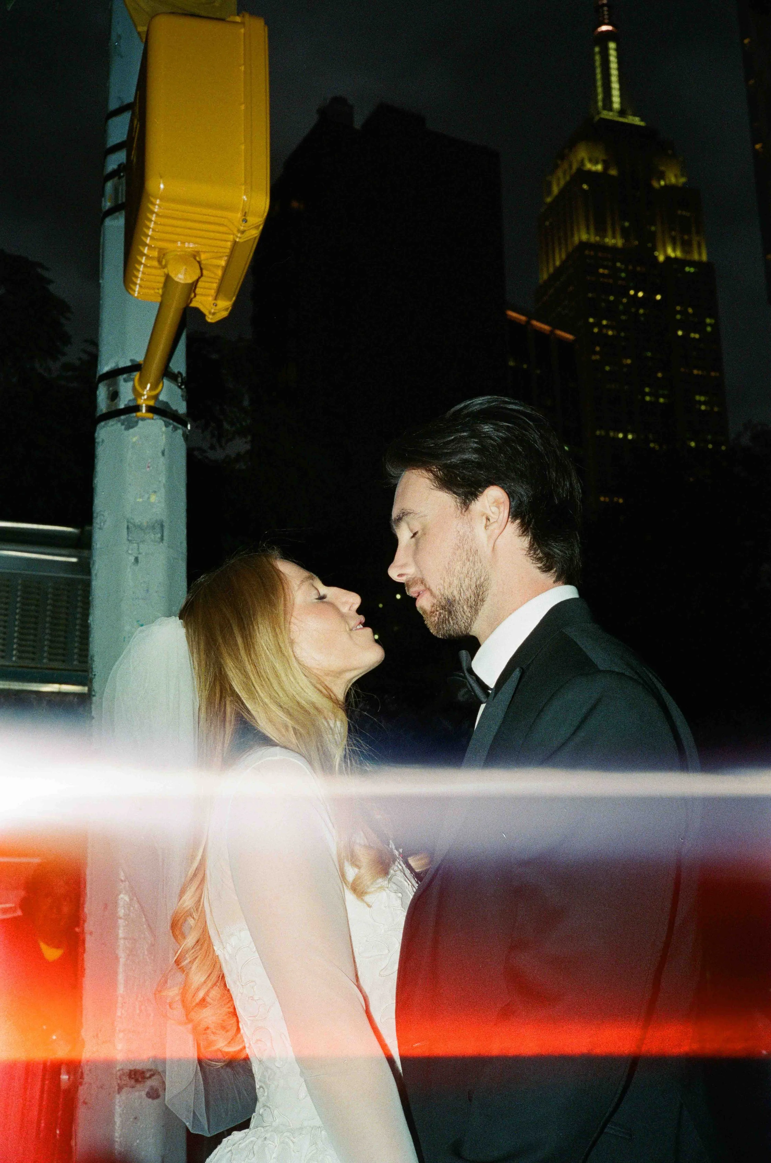 A bride and groom stand close together on a city street at night, with tall skyscrapers in the background, appearing to be about to kiss.