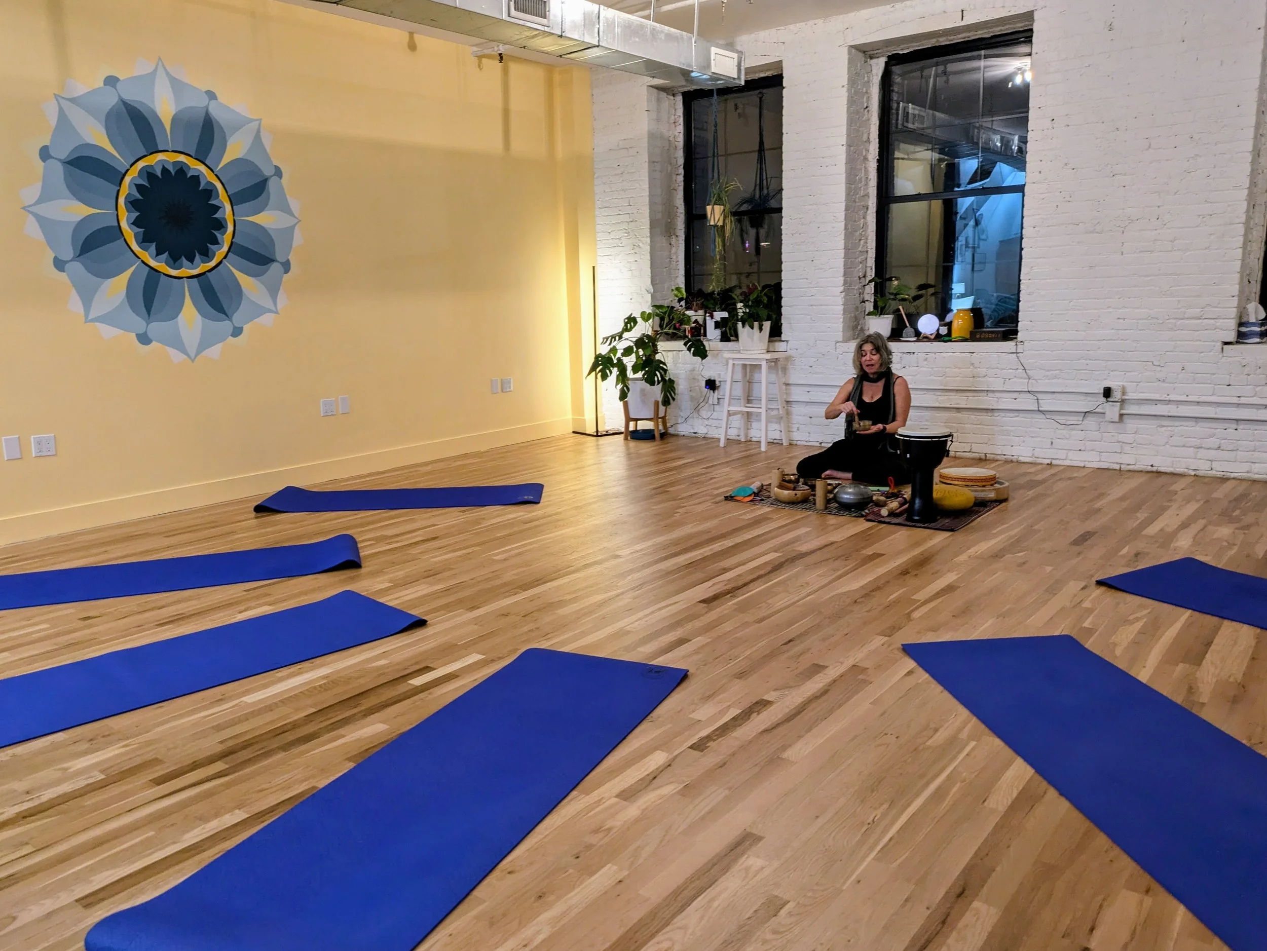 An empty yoga or meditation studio with five blue yoga mats on wooden floor; an instructor sitting on a rug near a white brick wall with large windows, surrounded by musical instruments and meditation tools.