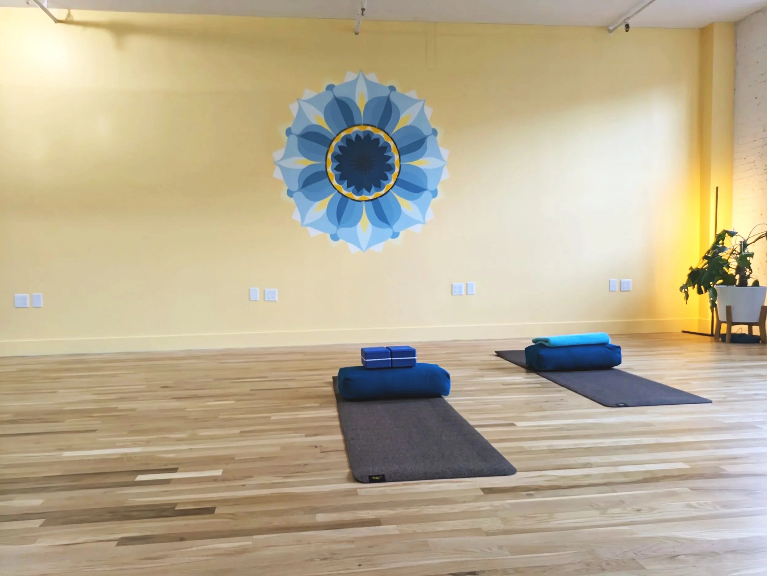 Yoga studio with two yoga mats on wooden floor, each with a rolled blanket, bolster, and folded towel. Yellow wall with a blue and white geometric mandala design, illuminated with warm lighting from the side, and a potted plant in the corner.