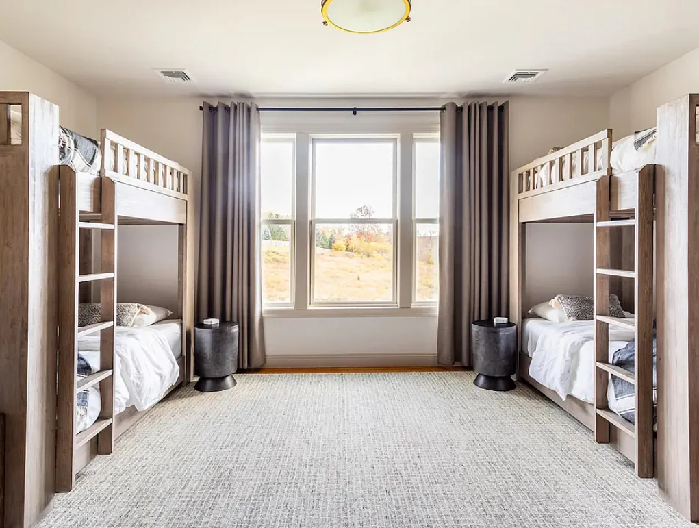 A bedroom with two wooden bunk beds, each with a ladder, positioned on either side of a large window with gray curtains and a view of an outdoor landscape.