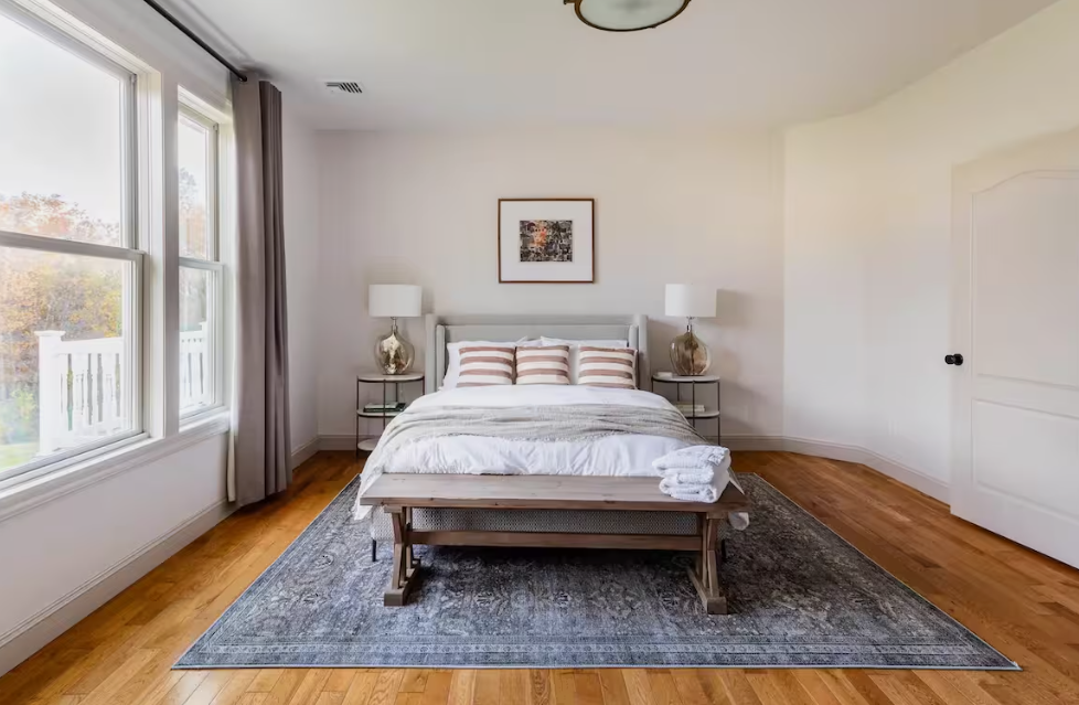 A neatly made bed with striped pillows in a bright bedroom with hardwood floors, two nightstands with lamps, a framed picture above the bed, and a large window with curtains.