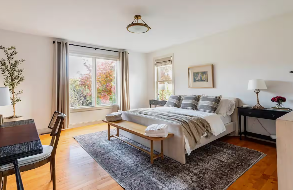 A bedroom with a large bed, two bedside tables with lamps, and a window showing trees with fall foliage.