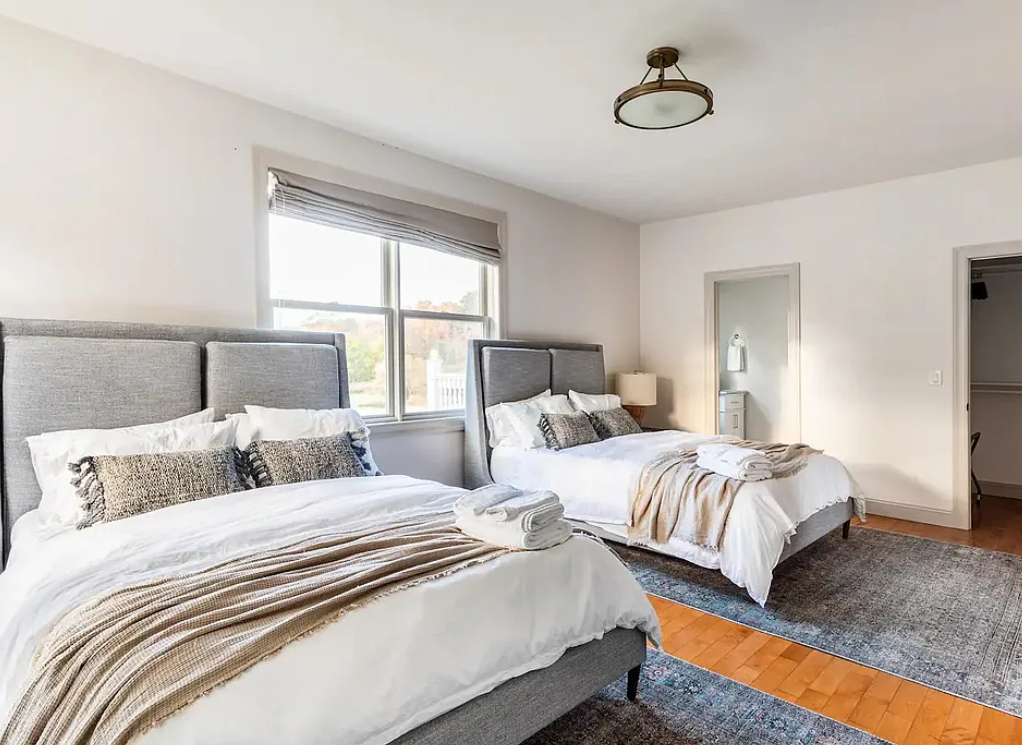 A bedroom with two large beds, each with gray upholstered headboards, white bedding, and various pillows. There are rolled towels on each bed, a window with blinds, a small nightstand with a lamp, and a door leading to another room or closet.