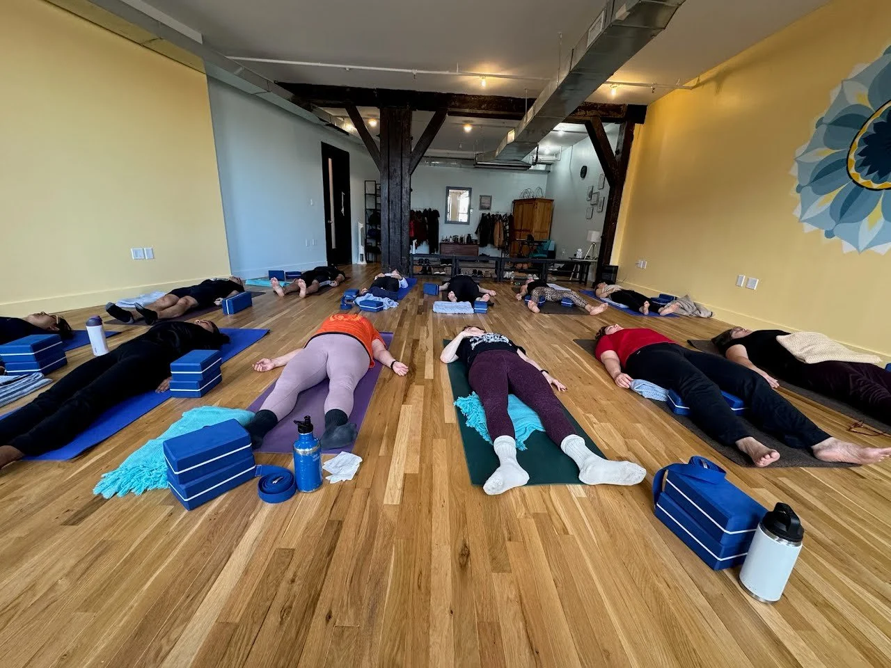 Yoga class in a studio with people lying on mats in relaxation pose, surrounded by yoga blocks and water bottles.