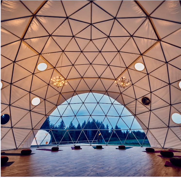 Interior view of a geodesic dome with wooden flooring, small round cushions, chandeliers, and large windows showing trees outside.