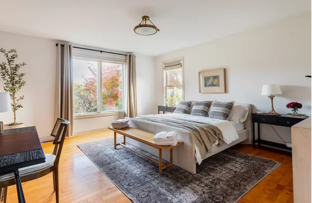 Bright bedroom with a large window, bed with pillows, bedside tables, and a decorative rug.