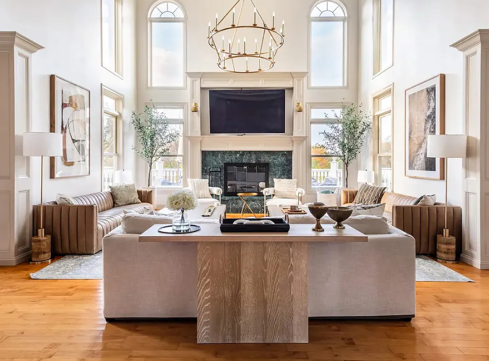 Bright living room with high ceilings, large windows, a chandelier, a fireplace, a wall-mounted TV, and modern furniture.