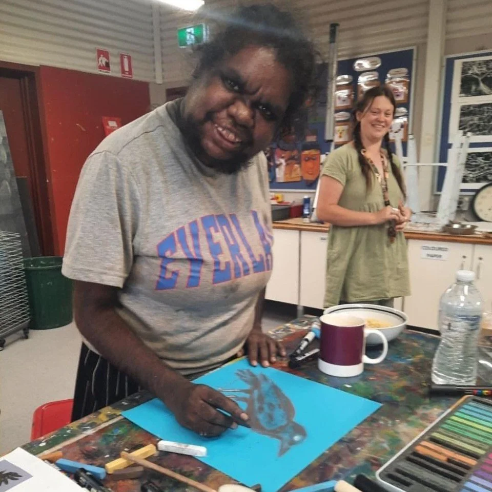 A man with curly hair and glasses drawing a colorful bird on blue paper with markers, in an art studio, smiling, with a woman in the background also smiling.