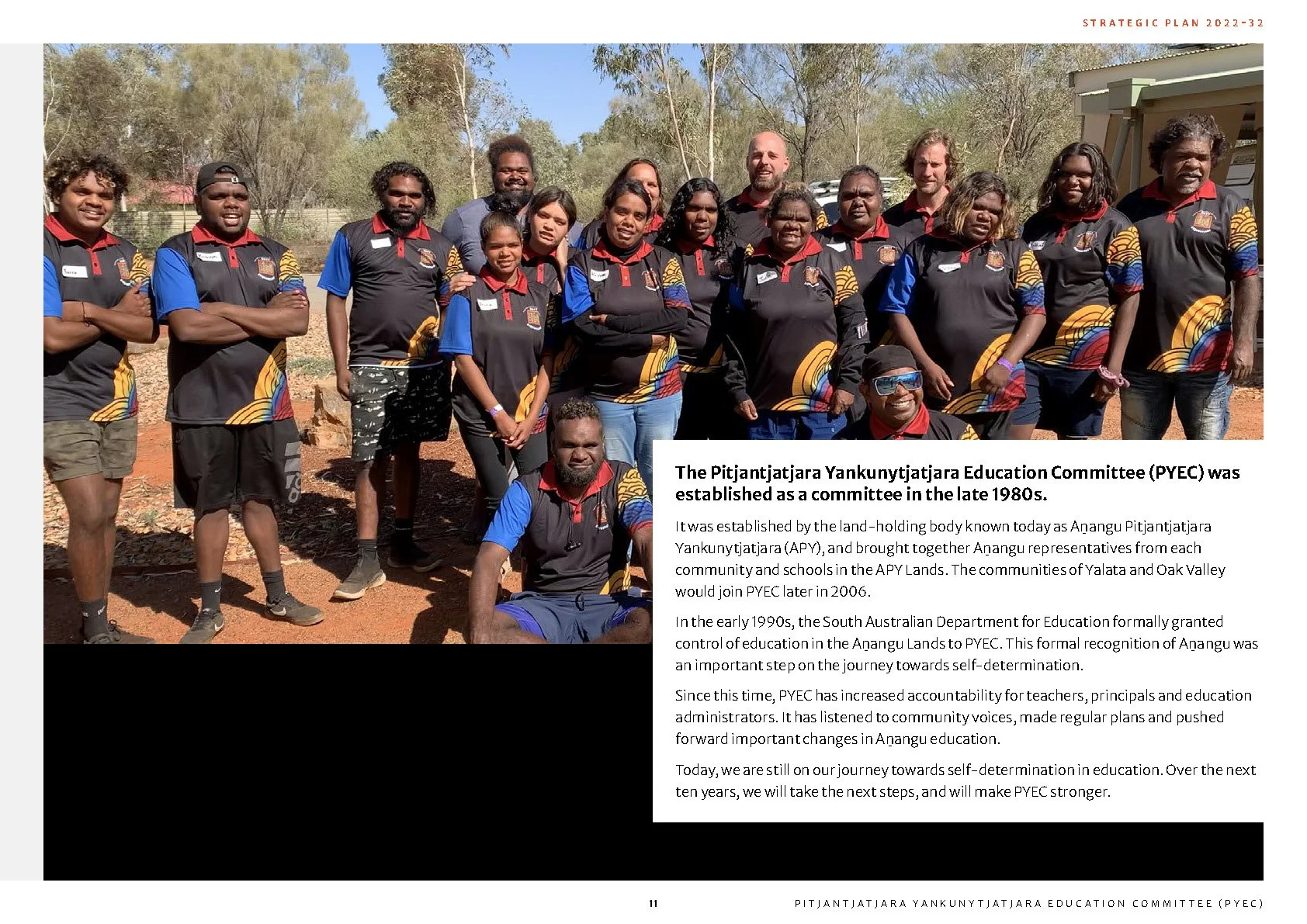Group of people wearing matching black and blue shirts with colorful patterns, standing outdoors in front of trees, with a document explaining the history and goals of the Pitjantjatjara Yankunytjatjara Education Committee (PYEC) and its role in educ