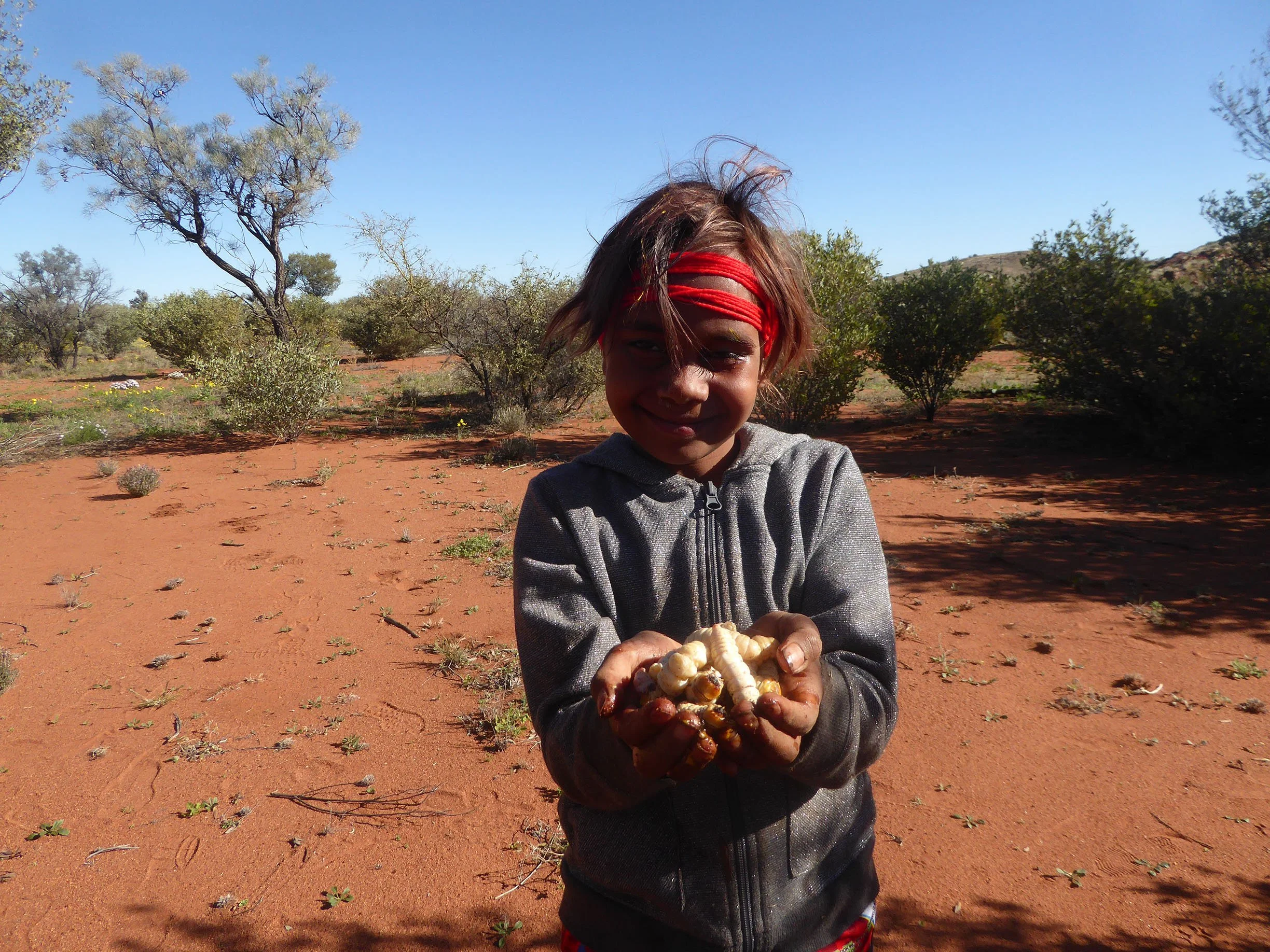 A young girl with dark hair, wearing a red headband and a gray zip-up hoodie, stands outdoors on red dirt ground, holding large white and brown fungi and smiling at the camera, with trees and bushes in the background under a clear blue sky.