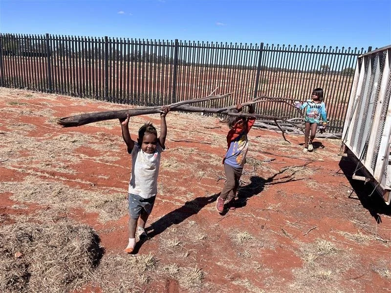 Three children holding a large tree branch outdoors on dry, reddish soil, with a black wooden fence and clear blue sky in the background.