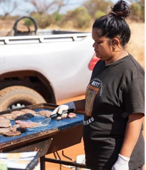 Woman grilling meat outdoors near a pickup truck on a sunny day.