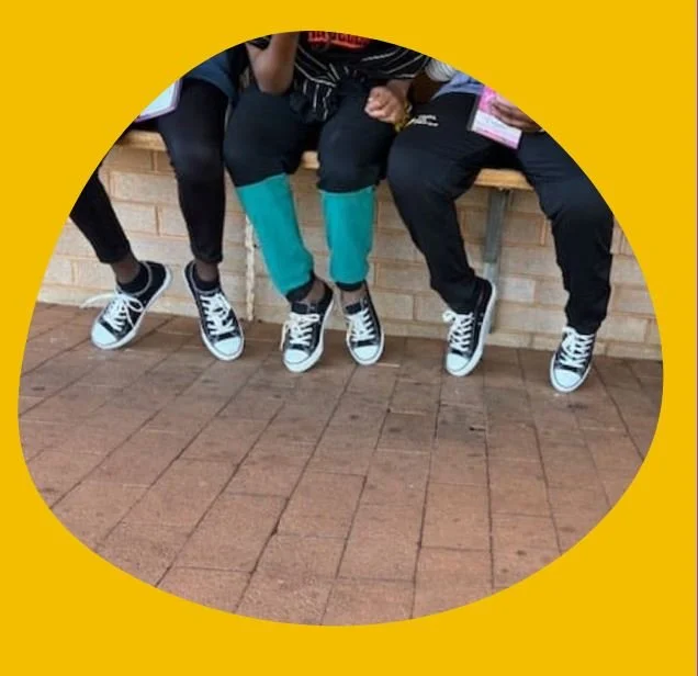 Four people sitting on a low brick wall with their legs hanging down, all wearing black and white sneakers, and blue or black pants, against a yellow background.