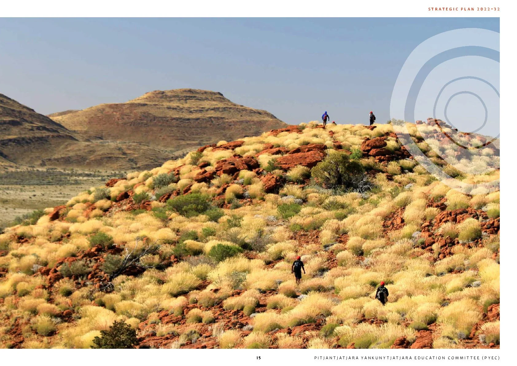 Hikers walking on a red rocky desert hill with yellow and green bushes, in a semi-arid landscape with distant hills and a clear sky.