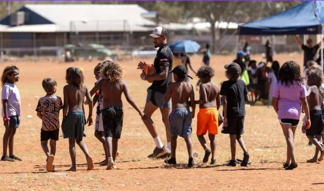 Children and an adult coach in a circle on a dry field during a sports practice or game.