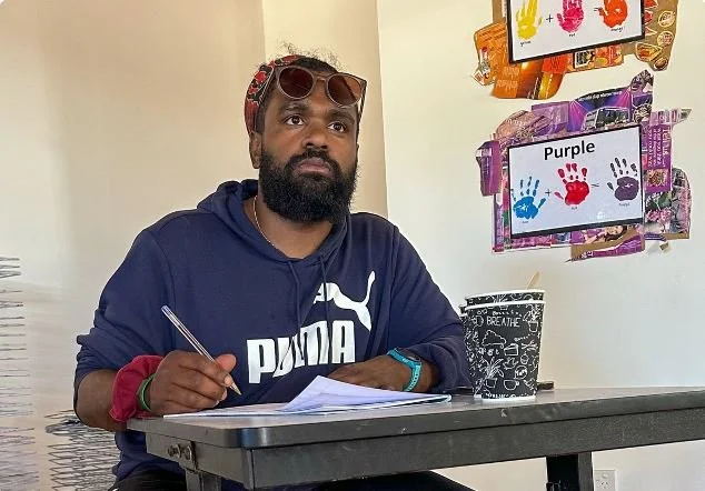 A man with sunglasses on his head, a beard, and a beard is sitting at a table with papers, holding a pen. Behind him, there are colorful handprint art projects including a purple handprint, on a wall.