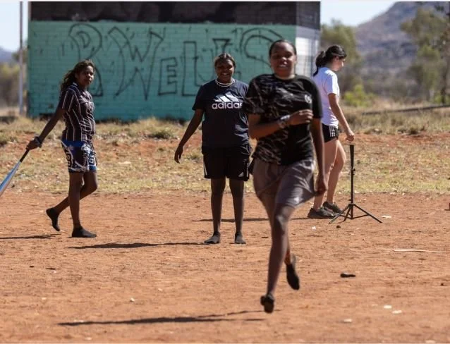 Four girls on a dirt field, with one running toward the camera, two standing and observing, and one walking with a microphone stand nearby, in an outdoor setting with a graffiti wall in the background.
