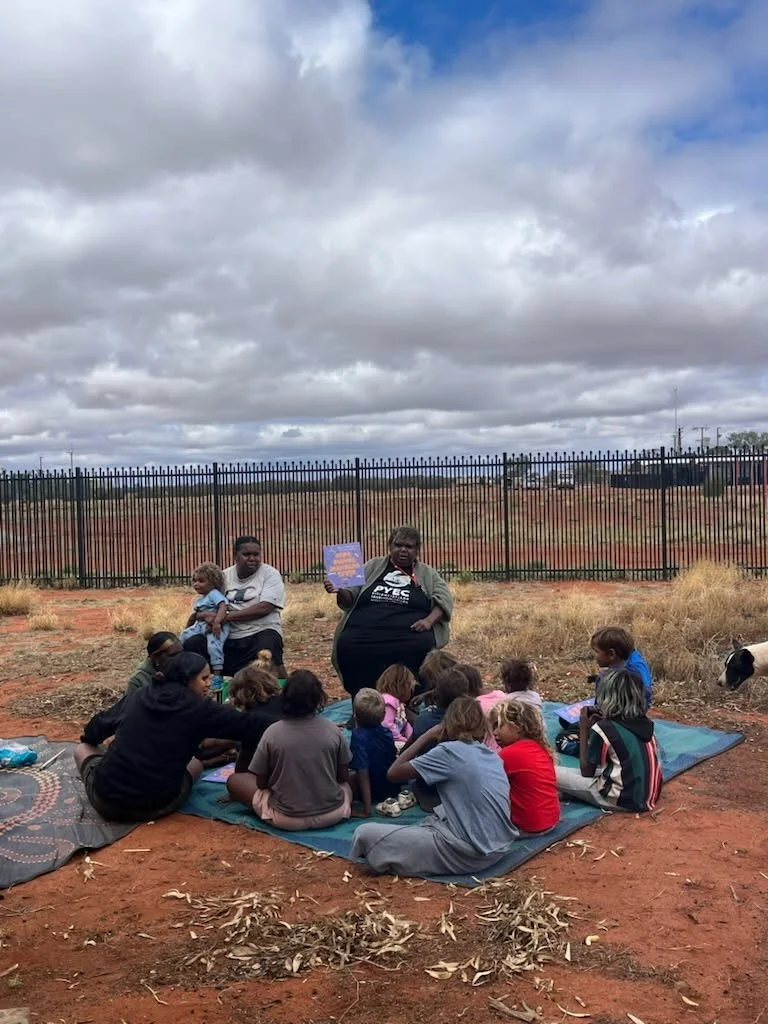 A group of children seated on a mat outdoors listening to a woman speaking, holding a colorful book. A man is sitting nearby holding a small child. The scene is set in a field with a black fence and cloudy sky overhead.