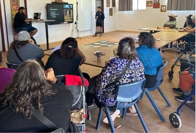 A group of people seated around a large rectangular table in a room, listening to a woman giving a presentation in front of a large screen with text and images. Some attendees have walkers nearby, and there is a podium and camera in the room.