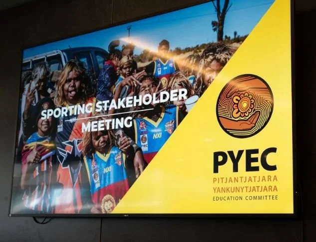 A large screen displays a sports stakeholder meeting with a group of children in sports uniforms and a yellow section with a logo and text for PYEY Education Committee.