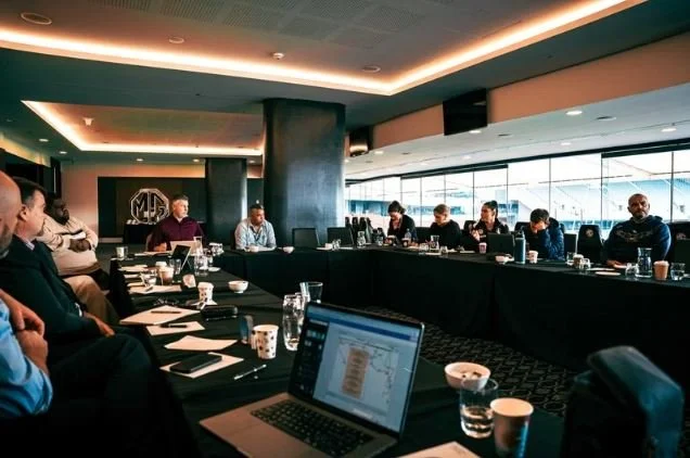 Business meeting in a conference room with attendees sitting around a U-shaped table, laptops, glasses of water, and coffee cups on the table, large windows in the background, and a MG logo on the wall.