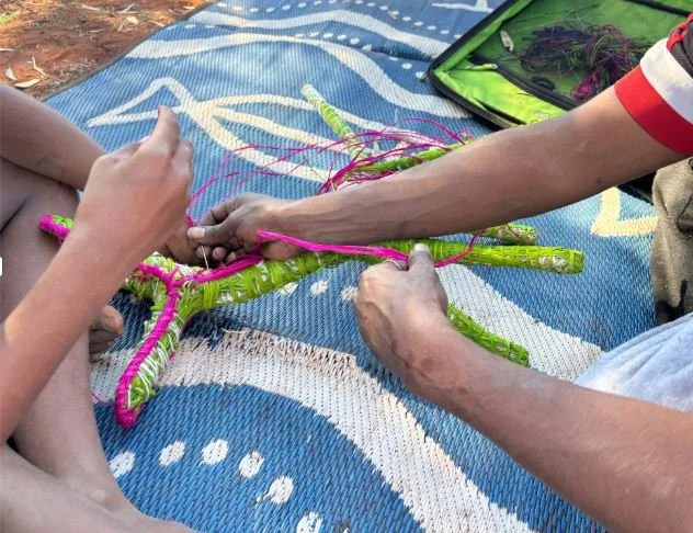Two people are weaving a colorful star-shaped craft using pink and green woven material on a blue outdoor mat, with a green bag nearby.
