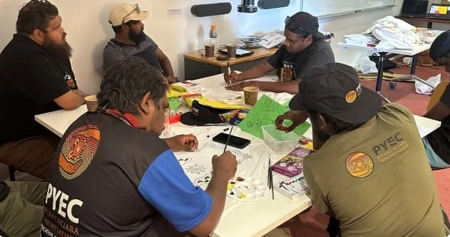 Group of people is sitting around a table engaged in a casual meeting or game, with papers, pens, snacks, and drinks on the table.