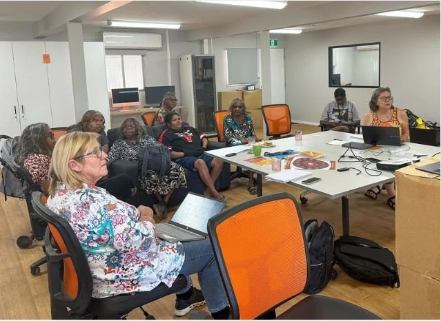A diverse group of women attending a meeting or workshop in a conference room with a long table, laptops, and papers.