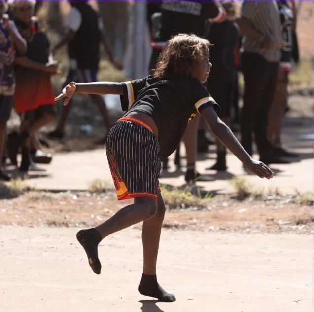 Young girl throwing a stone in a dirt area with onlookers in the background.
