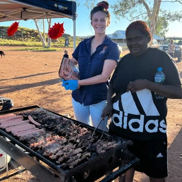 Two women standing at an outdoor barbecue grill with skewered meat, in a park-like setting with a tent decorated with red pom-pom ornaments, and other people and trees in the background.