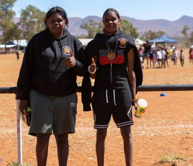Two young girls standing outdoors in front of a fence, wearing black hoodies and shorts, giving thumbs up. The background shows a group of people and mountainous terrain.
